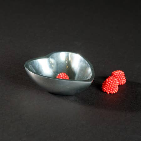 A shiny aluminum heart-shaped bowl sits on a dark surface, holding one bright red bumpy candy. Two identical candies lie beside it, creating a vivid contrast with the metallic home decor centerpiece and its dark backdrop.