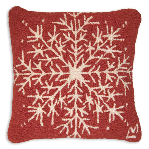 The Flake Pillow features a square plush design with a textured surface, showcasing an intricate hand-hooked snowflake pattern crafted in wool on a deep red background. The detailed, symmetrical pattern of its multiple branching arms brings a cozy wintry feel to the item.