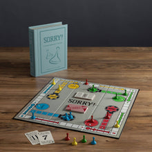 Load image into Gallery viewer, The image showcases the &quot;Sorry! Vintage Bookshelf Edition&quot; game board on a wooden table. The open board displays colorful pawns and cards, while the closed, collectible box resembling a fabric-wrapped book stands upright in the background.
