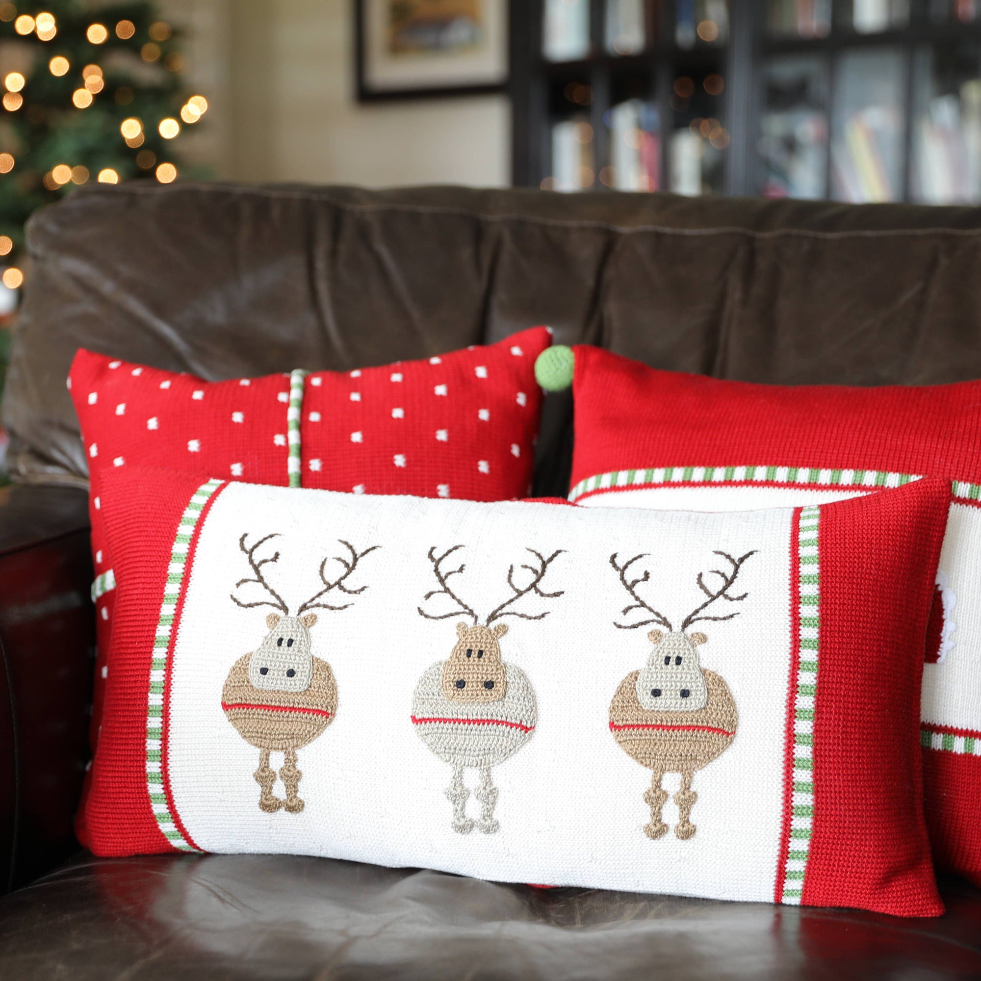 A cozy holiday scene features a festive brown leather couch adorned with three handmade Round Reindeer Lumbar Pillows. The pillows exhibit charming reindeer designs and come in vibrant red with white polka dots and green accents. A softly lit Christmas tree and an unfocused bookshelf enhance the warm, festive ambiance in the background.