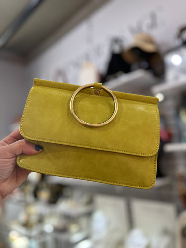 In a stylish store, someone holds the Aria Ring Bag - Spring/Summer, featuring vegan leather and a large gold ring clasp. Blurred shelves of accessories and hats form the background while the bag's sleek design and smooth texture take center stage.