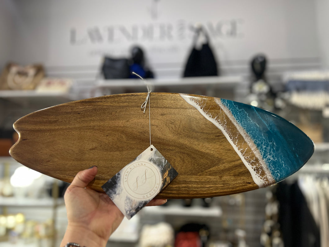 A hand holds an Acacia Resin Surfboard in a store, which is crafted from Acacia hardwood. The wall art piece features a realistic ocean wave crashing onto a sandy beach, made with eco-friendly resin. A Lavender & Sage tag is attached to the board. Shelves with various items, including bags and decor, are visible in the background.