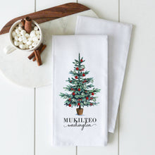 Load image into Gallery viewer, From our custom-designed kitchen collection, the Tea Towels - Summit in white showcase a decorated Christmas tree adorned with red and silver ornaments. Below the tree, &quot;Mukilteo, Washington&quot; is elegantly scripted. The towel is beautifully displayed on a white marble cutting board alongside marshmallows and cinnamon sticks on a wooden surface.
