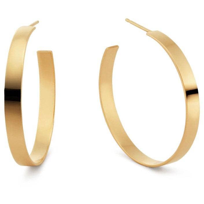 The Carrie Flat Hoops, a pair of 18k gold plated earrings, glimmer against a plain white background. These wide, tarnish-resistant earrings feature a smooth, polished surface; one gracefully faces forward while the other is slightly angled to showcase their elegant circular design.