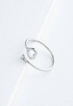 Load image into Gallery viewer, The Ada Silver Heart Ring, a minimalist accessory crafted from stainless steel in the shape of an open heart, sits elegantly on a white wooden surface. Its adjustable band boasts a seamless and sleek design with a shiny finish, offering a modern and elegant look. This chic piece is perfect for adding everyday elegance to any outfit.
