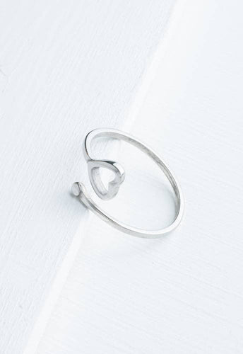 The Ada Silver Heart Ring, a minimalist accessory crafted from stainless steel in the shape of an open heart, sits elegantly on a white wooden surface. Its adjustable band boasts a seamless and sleek design with a shiny finish, offering a modern and elegant look. This chic piece is perfect for adding everyday elegance to any outfit.