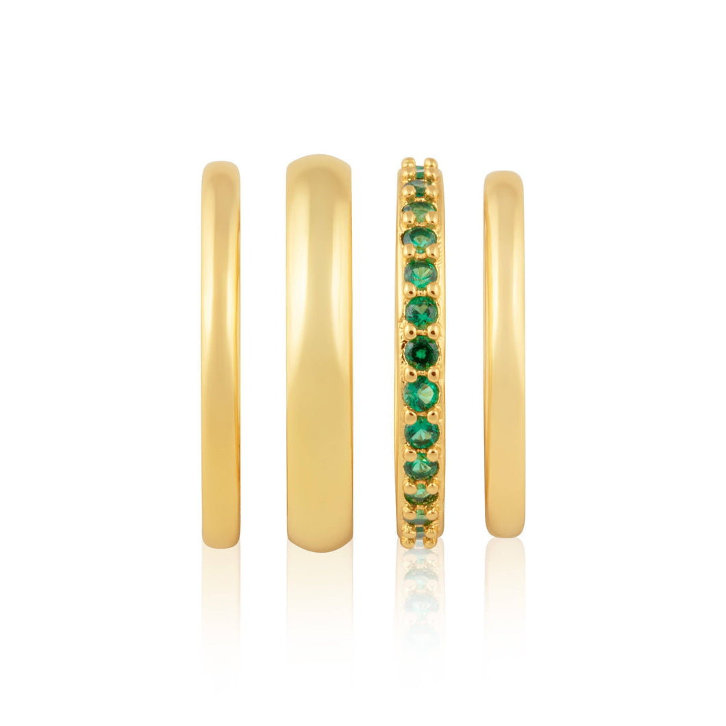 The Faye Emerald Stacked Bands are showcased in a vertical arrangement, featuring four gold rings from left to right: an 18k gold plated band, a slightly wider stainless steel ring, a band encrusted with green cubic zirconia, and another plain polished gold band. Each ring has a reflective surface that creates a mirrored effect below.