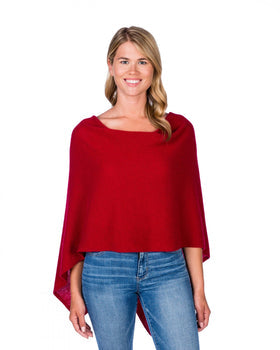 A smiling woman with long blond hair models the Cashmere Poncho, a vibrant red poncho-style top that serves as an ideal dress topper over blue jeans. The one-size piece features a wide neckline and drapes loosely. She stands against a plain white background, gazing directly at the camera with her arms relaxed at her sides.