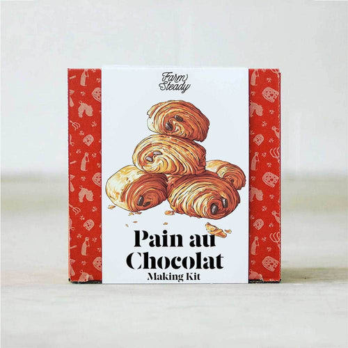 The Pain au Chocolat Making Kit's packaging features a bold design in vibrant red, decorated with images of white kitchen utensils and motifs inspired by French pastries. The box is highlighted by an illustration of five neatly stacked golden-brown, flaky Pain au Chocolat. The product name stands out in bold black lettering.