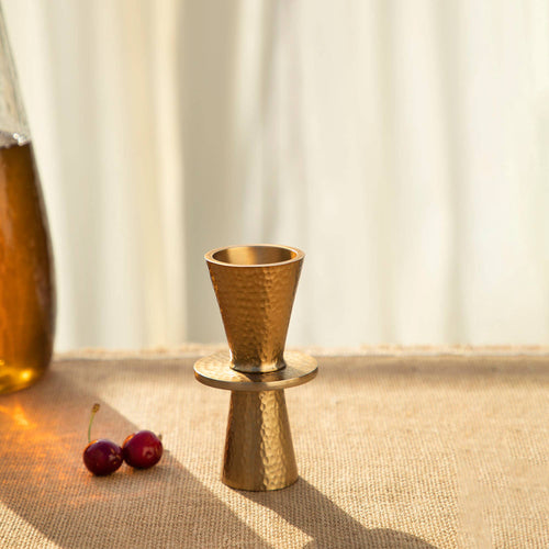 A jigger with a hammered brass design is positioned on a woven surface, making it an ideal addition to any home bar. It features two cone shapes joined at their wide ends, casting a shadow to the right. Nearby, two cherries sit in the softly lit setting, with part of a glass bottle faintly visible on the left in the blurred background.