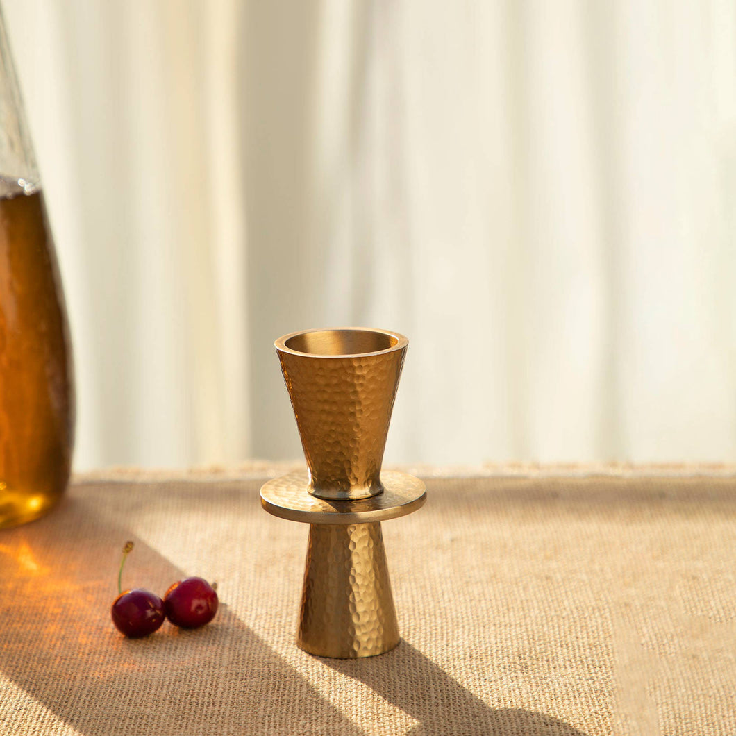 A jigger with a hammered brass design is positioned on a woven surface, making it an ideal addition to any home bar. It features two cone shapes joined at their wide ends, casting a shadow to the right. Nearby, two cherries sit in the softly lit setting, with part of a glass bottle faintly visible on the left in the blurred background.