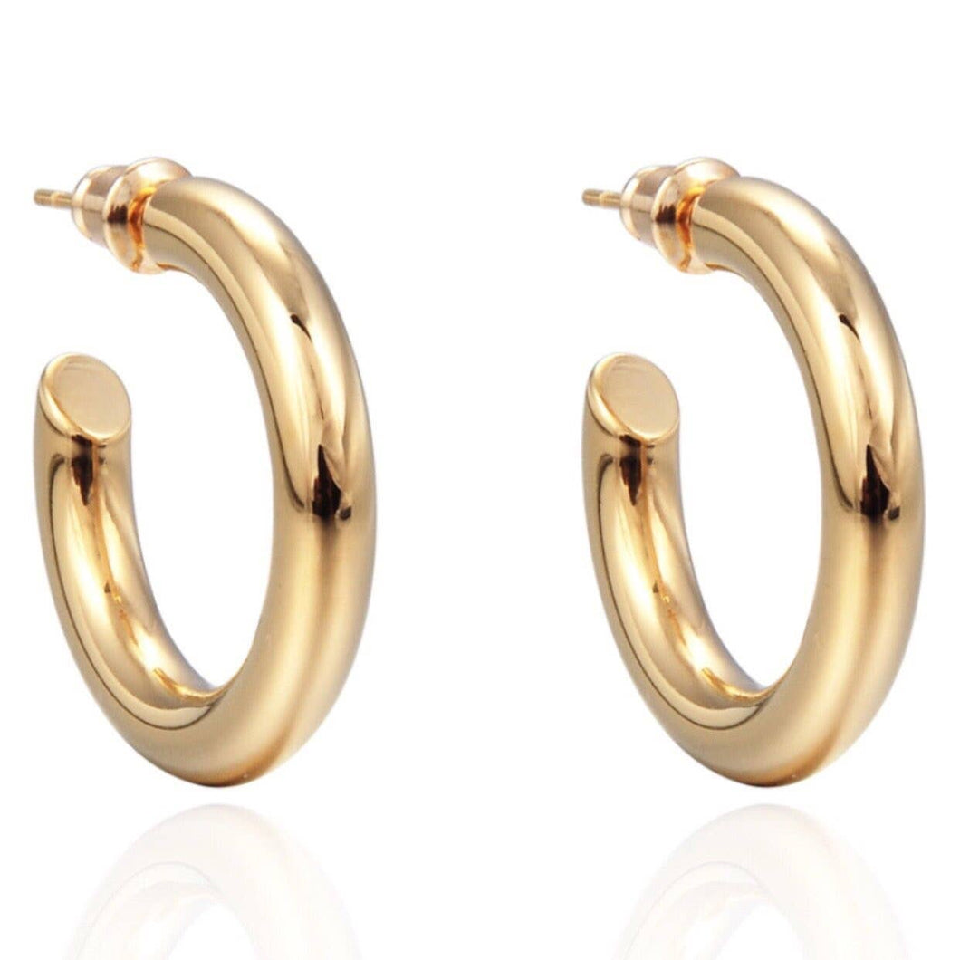 The Anya Hoop earrings, a pair of shiny 18K gold-plated hoop earrings, are displayed against a white background. These midi-sized earrings are thick and smoothly polished, each featuring a straight post for pierced ears. Their hypoallergenic design showcases a reflective surface that catches the light, highlighting their glossy finish.