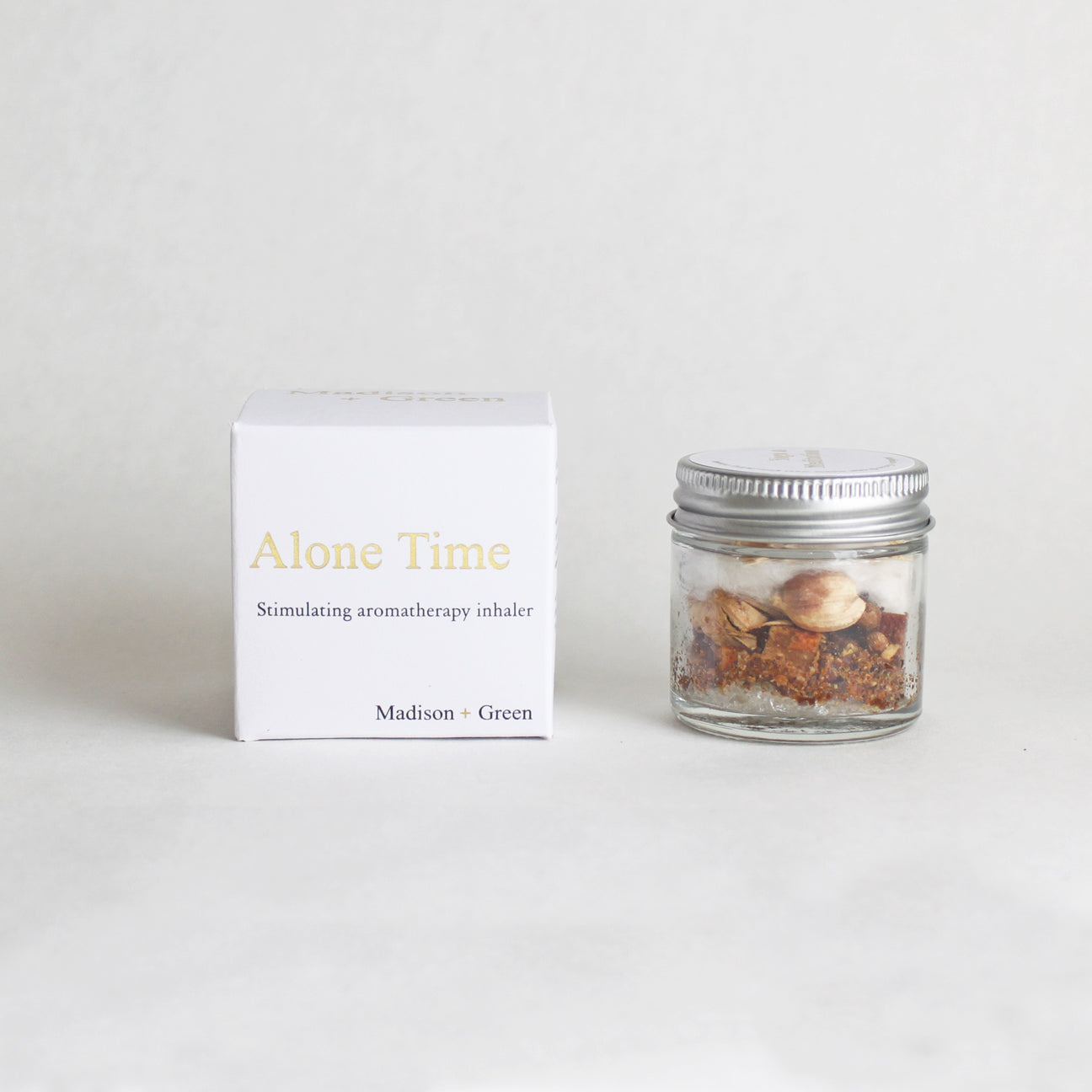 A small, clear glass jar with a silver lid holds dried herbs and spices, positioned next to a white box labeled "Alone Time: Stress Relief aromatherapy inhaler" in gold and gray text from the brand "Madison + Green." The product belongs to the "Aromatherapy Inhalers - 13 varieties" collection. The background is simply white.