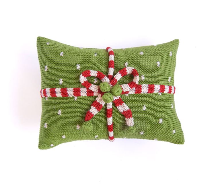 Introducing the Green Gift Mini Pillow: a stunning example of Armenian craftsmanship. This rectangular, green knit pillow is adorned with small white dots and features a red and white striped knitted bow wrapped around it like a festive gift ribbon. By choosing this beautiful piece, you're also supporting fair trade practices.