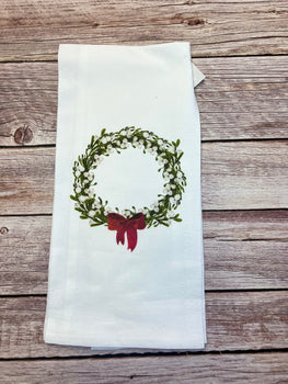 As part of our Holiday Collections, the Winter/Christmas Tea Towels feature a handcrafted green wreath design adorned with delicate white berries and a charming red bow. Elegantly displayed on a rustic wooden surface, this white tea towel adds a festive touch to any kitchen setting.