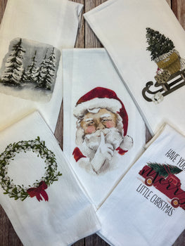 Five tea towels from the Winter/Christmas collection are showcased in the Holiday Collections. The designs feature a snowy pine tree scene, Santa Claus with a finger to his lips, a Christmas tree on a sled, a holiday wreath adorned with a red bow, and the phrase 