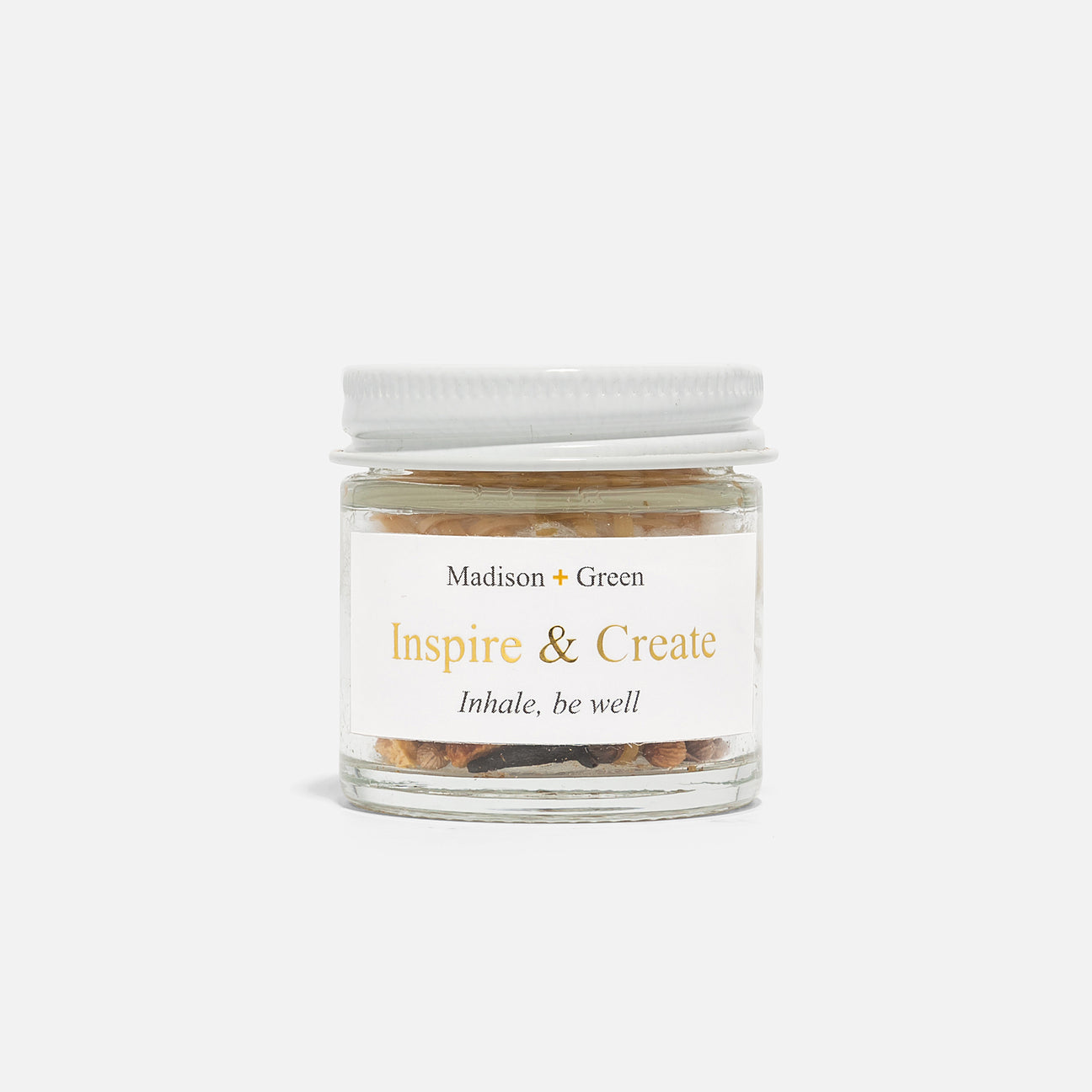 A petite glass jar featuring a white lid houses natural stones and serves as an aromatherapy inhaler. The label displays "Madison + Green: Inspire & Create" with the phrase "Inhale, be well" beneath it. This product, part of the Aromatherapy Inhalers collection available in 13 varieties, is perfect for stress relief and adds a calming essence to any simple white setting.