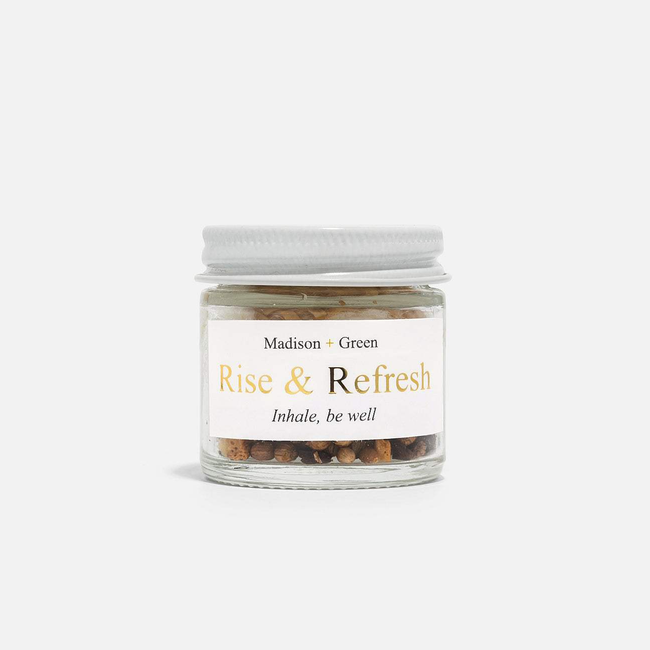 A small glass jar with a white lid holds a blend of stress-relieving aromatherapy beads. The label features &quot;Madison + Green&quot; at the top, &quot;Rise &amp; Refresh&quot; prominently in the center, and &quot;Inhale, be well&quot; at the bottom. Made with organic essential oils, it&#39;s presented on a plain white background as part of their Aromatherapy Inhalers line, which includes 13 different varieties.