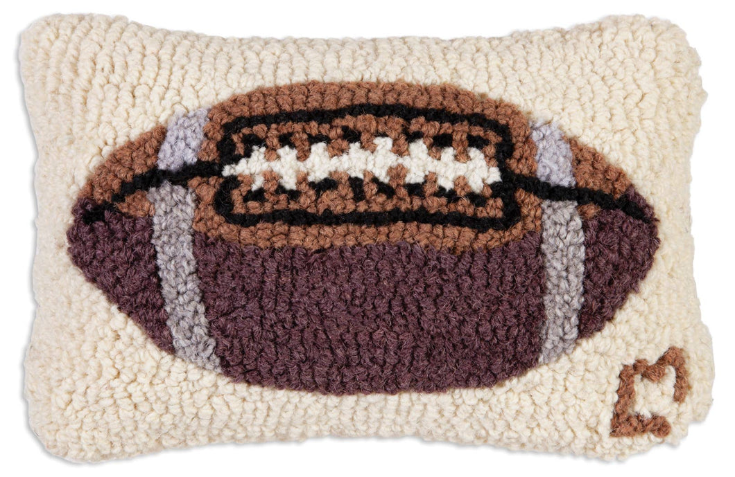 The Football Pillow is a small, rectangular cushion with a tufted texture, showcasing a football motif at its center. It is hand-hooked with natural wool to provide realistic stitching details. This cream-colored pillow is handcrafted in India and features an adorable heart design in the bottom right corner.