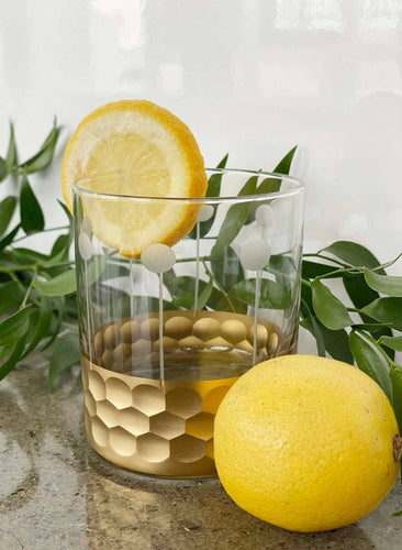 A Moroccan Gold Hammered Glass from the set of four sits elegantly on the marble, featuring a honeycomb pattern and adorned with a slice of lemon on its rim. Alongside it, a whole lemon enhances the display, while green leaves form a lush backdrop. The glass's opaque dots add an extra layer of charm to its sophisticated design.