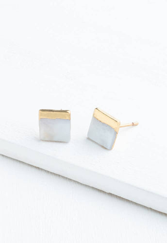 The Kelly Mother of Pearl Stud Earrings, featuring a minimalist design with square studs and 18k gold-plated borders, are showcased on a light wooden surface. These hypoallergenic earrings have a smooth finish that offers a subtle yet elegant look.