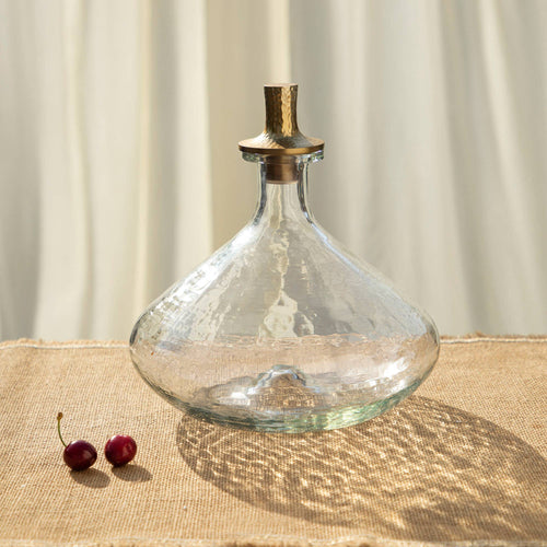 The Decanter Wide, featuring a clear pebbled glass design and a gold-finished stopper, rests on a textured beige surface, casting intricate shadows. Nearby, two cherries add to the scene while softly blurred light-colored drapes in the background create a warm, elegant ambiance.