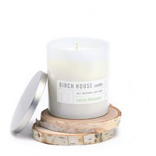 Load image into Gallery viewer, A white frosted glass Cactus Blossom Candle by Birch House Candles, made from all-natural soy wax, is lit on round birch wood slices with a silver lid beside it. The minimalist backdrop enhances its elegance.
