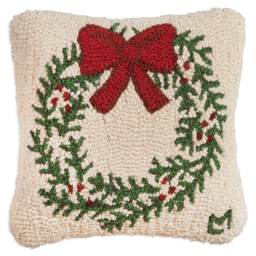 The Christmas Wreath Pillow is a handcrafted wool cushion that showcases a hooked texture with an exquisite design of a Christmas wreath. The vibrant wreath, adorned with green leaves, red berries, and a large red bow, is set against a cream background to enhance the festive spirit. It is proudly crafted in accordance with Fair Labor Association standards.