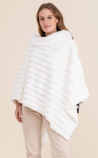 A person wearing a Faux Fur Poncho in white with horizontal stripes stands in front of a neutral background. With long, light brown hair and hoop earrings, they are dressed in light-colored pants and exude stylish warmth as their hands hang relaxed beside them.