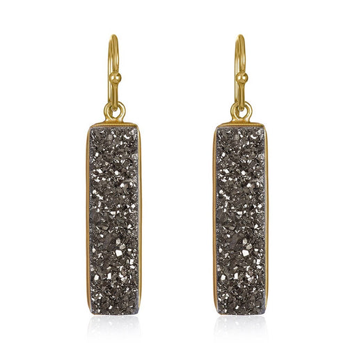 The Druzy Baguette Earrings: Gold-Filled feature a pair of rectangular drop earrings adorned with textured dark gray druzy stones set within a gold-plated frame. These earrings gracefully dangle from straightforward gold hooks, highlighting a sparkling, crystalline surface that contrasts beautifully with the smooth metallic edges.