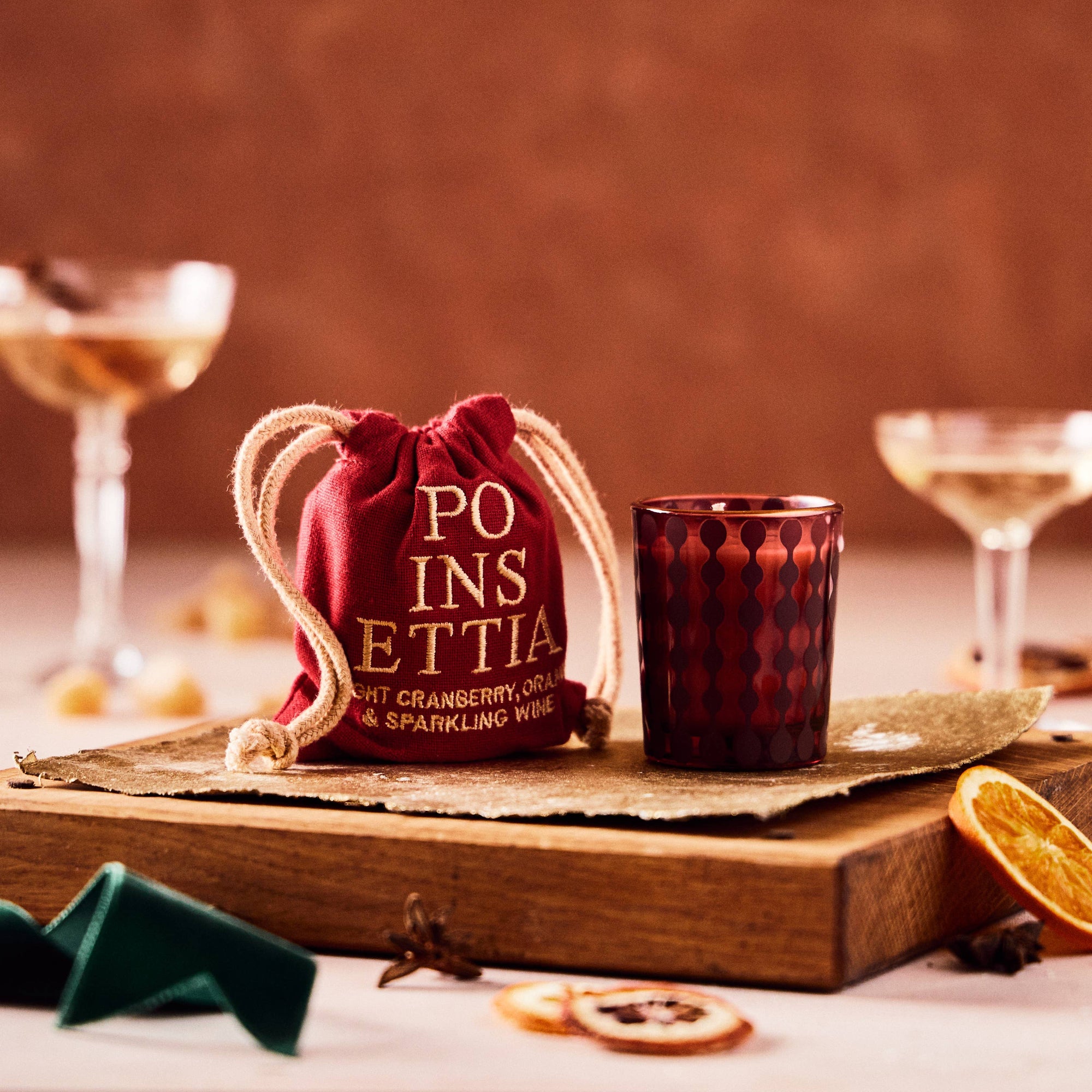 A red Poinsettia Candle 2.5 oz sits on a wooden tray next to a drawstring bag labeled "Poinsettia." In the background, two elegant cocktail glasses filled with a Poinsettia cocktail, showcasing cranberry hues and sparkling wine, stand against a warm brown wall. Dried orange slices and decorative items are scattered around the scene.