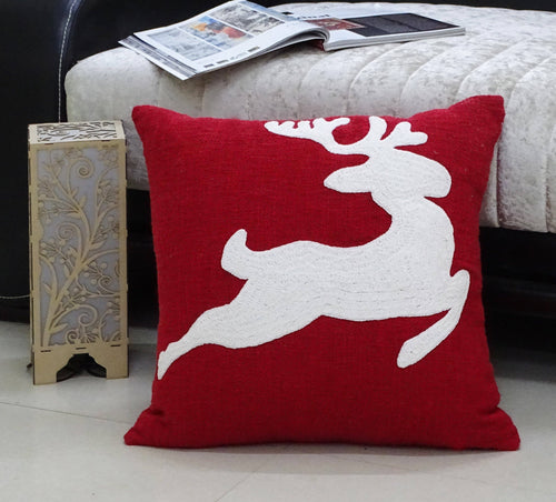 A Reindeer Throw Pillow, featuring a festive red color with a white silhouette of a leaping reindeer, rests on the floor. Beside it is an ornate wooden lantern adorned with a floral design. In the background, an open magazine is laid out on the light-colored sofa, while the floor remains smooth and light.