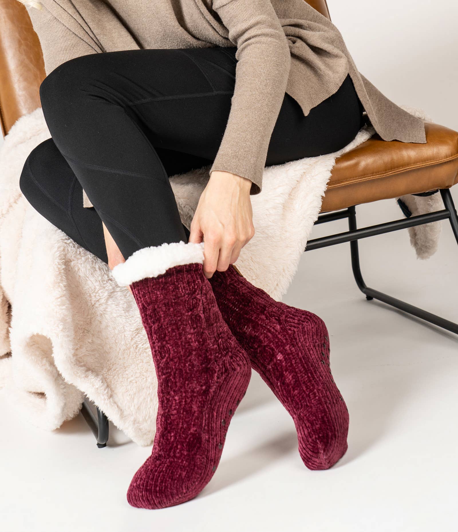 A person sits on a brown chair, wearing black leggings and a beige sweater. They pull on a pair of Beyond Soft Slipper Socks in maroon chenille with white fleece lining. A soft, cream-colored blanket drapes over the chair, creating a cozy atmosphere with non-slip soles for added comfort.