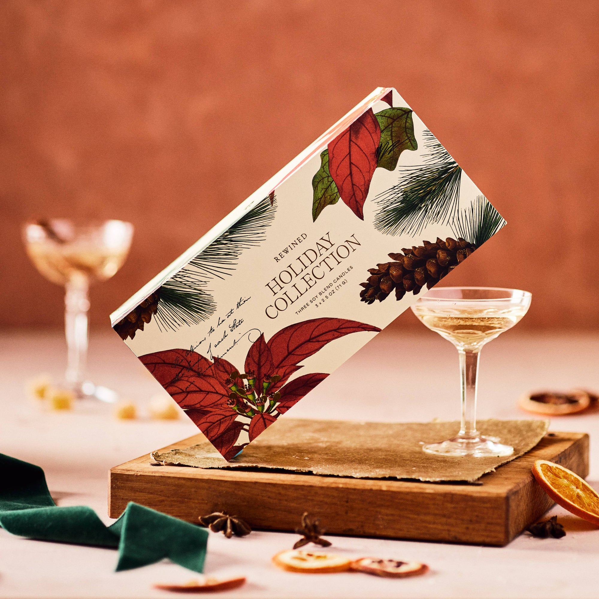 A festive gift box labeled "Holiday Candle Gift Set" is decorated with illustrations of red poinsettias, green leaves, and a pinecone. Designed for the holiday season, it's propped up on a wooden block beside a filled champagne coupe. Dried orange slices and star anise add a fragrant touch to the scene.